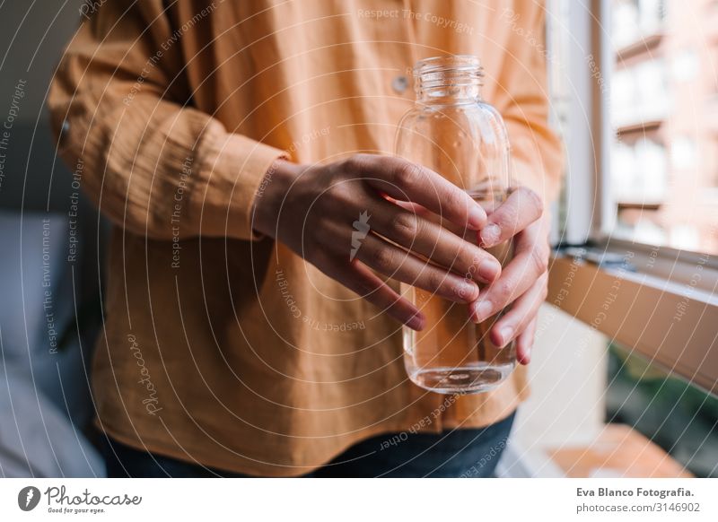 junge schöne Frau am Fenster zu Hause, die Wasser trinkt. Lebensstil Glas Flasche trinken Becher Tasse gelb Liebe Umarmen heimwärts niedlich Tag traumhaft