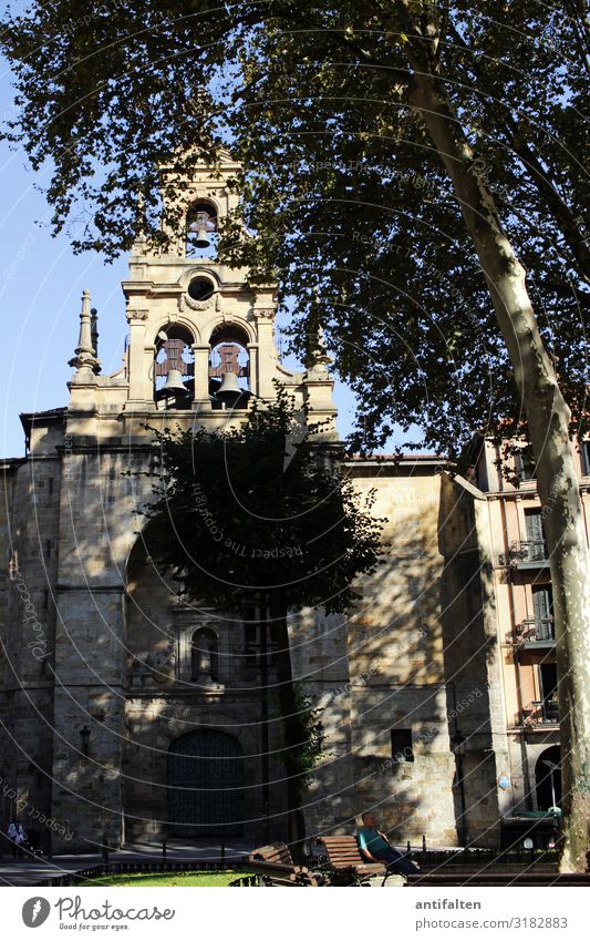 Ein Mann sitzt im Schatten auf einer Parkbank an einem Kirchplatz Kirche Bäume Platz Glockenturm Bilbao Sommer Sonne Bank Pause Erholung Außenaufnahme ruhig