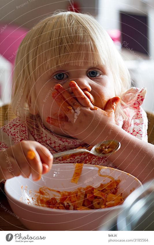 Kleines, blondes, süßes Mädchen ist Nudeln mit Tomatensoße und verschmiert mit der Hand ihr Gesicht mit roter Sauce, sie hält den kleinen Löffel in der Hand, über einer Schale und schaut neugierig in die Kamera, zuhause in der Wohnung.