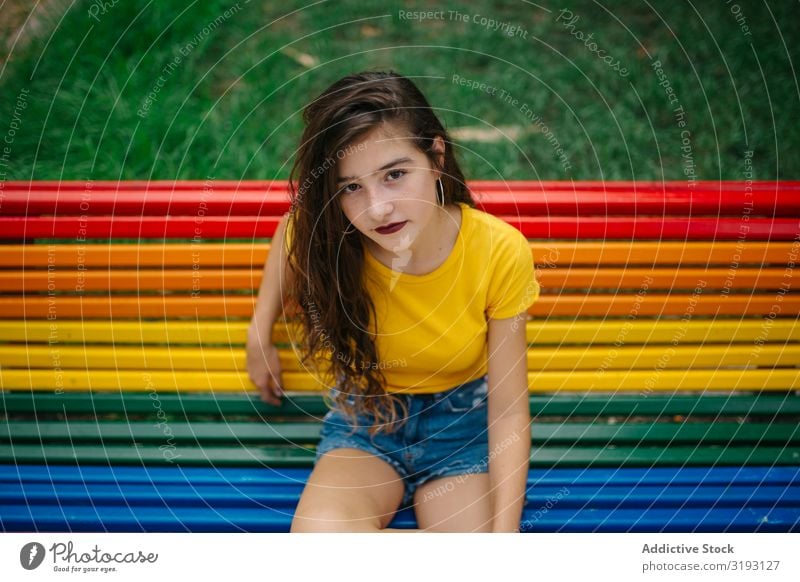 Fröhliche Frau auf Regenbogenbank sitzend Bank Park Lächeln Blick in die Kamera lässig Lifestyle Freizeit & Hobby Jugendliche ruhen Erholung Sommer Glück Freude