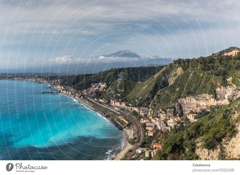 Blick aus der Vogelpespektive auf eine Felsige, eng bebaute Bucht. Eine Schienentrasse schlängelt sich am Meer entlang, am Horizont taucht der Ätna aus der Wolkendecke