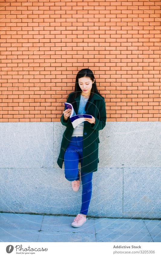 Asiatischer Student liest Lehrbuch in der Nähe der Ziegelmauer Schüler lesen Handbuch anlehnen Wand Backstein asiatisch Frau Studium Jugendliche Notizbuch