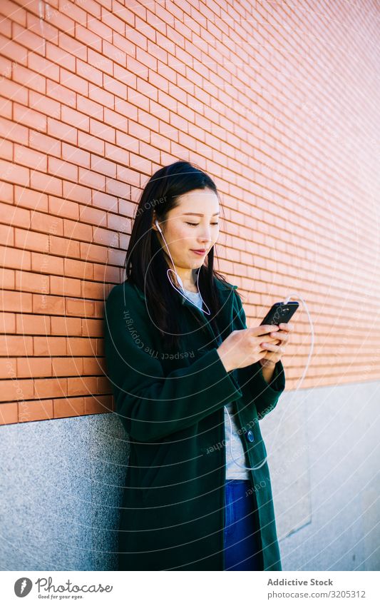 Asiatische Frau hört Musik in der Nähe einer Ziegelmauer hören PDA benutzend Wand anlehnen Backstein Straße Großstadt asiatisch urwüchsig Lifestyle