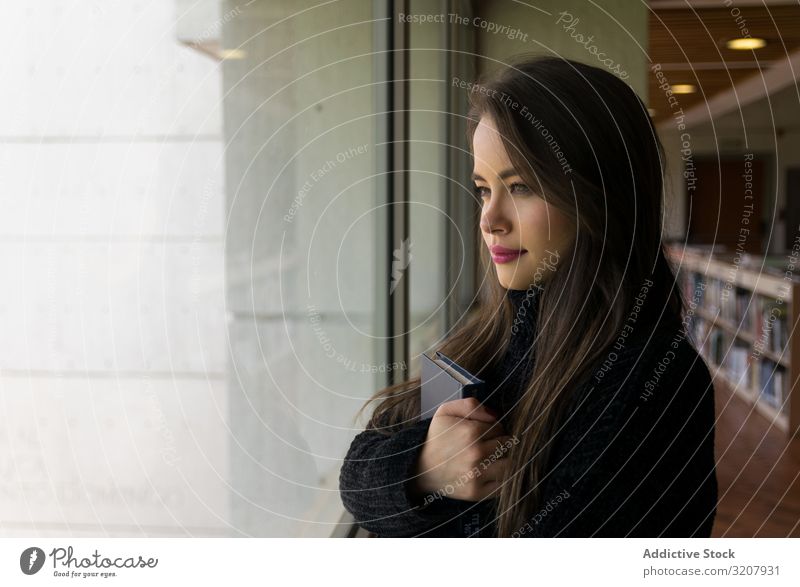 Hübsche Frau mit einem Buch, das aus dem Fenster schaut attraktiv klug clever schön brünett jung niedlich Schönheit hübsch heiter lässig Schüler elegant