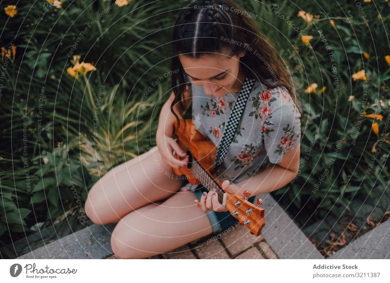 Fröhliche moderne Frau spielt Ukulele Großstadt Lächeln lässig sitzen Straßenbelag Blumenbeet trendy lange Haare T-Shirt zufrieden Instrument jung Spaß Freizeit