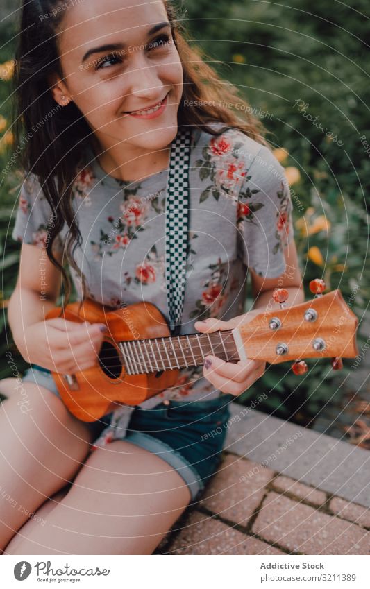 Fröhliche moderne Frau spielt Ukulele Großstadt Lächeln lässig sitzen Straßenbelag Blumenbeet trendy lange Haare T-Shirt zufrieden Instrument jung Spaß Freizeit