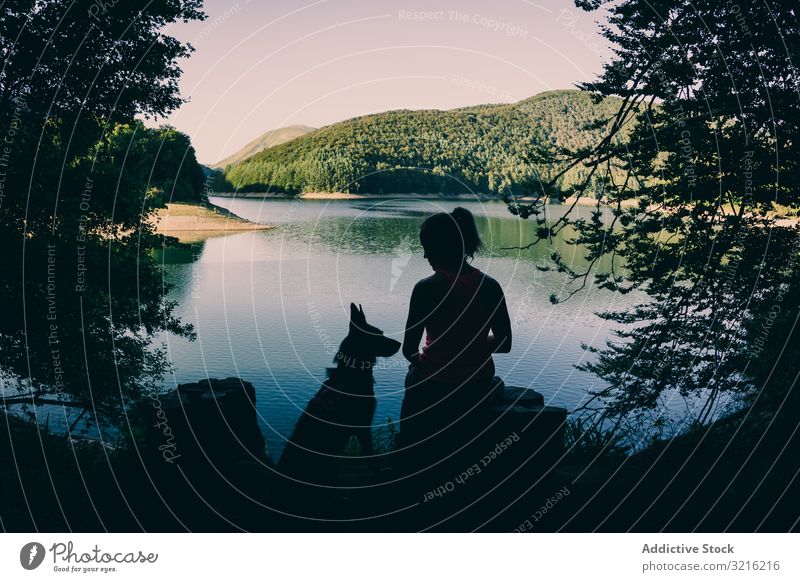 Frau mit Hund ruht sich am schönen See aus Silhouette aussruhen bewundernd Reisender Tourismus wandern malerisch Wald Sommer Tag Haustier Tier Freundschaft