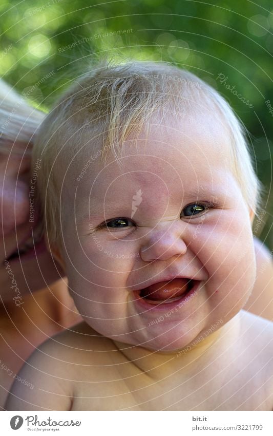 Kleiner, süßer Wonneproppen lacht, quitschend in die Kamera. Glückliches Baby, Kleinkind mit Mutter lacht bis zu den Ohren, draussen in der Natur. Blondes Mädchen im Schwimmbad mit fröhlicher Mimik, Gesichtsausdruck. Lichtpunkte der grünen Wiese hinten.