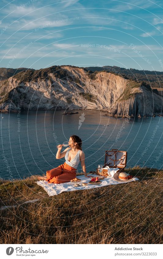 Frau mit Glas Getränk auf Picknickmatte mit Blick auf Meer und Berge Seeküste Strand Sommer lesen Freizeit Himmel MEER sich[Akk] entspannen trinken Urlaub