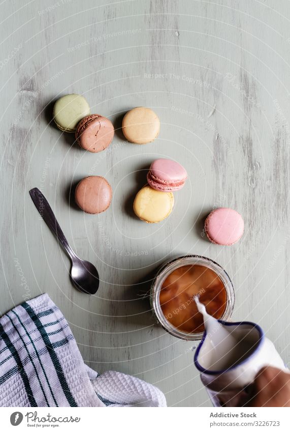 Erntehelfer, der in der Nähe von Makronen Milch zum Kaffee gibt Frühstück eingießen melken Tasse Löffel Serviette Tisch Lebensmittel Morgen Molkerei Mahlzeit