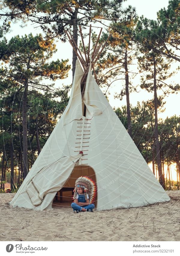 Zufriedenes Kind mit authentischem Hut im Zelt Junge Wigwam Inder Feder Inhalt erfreut Latzhose Jeansstoff Stehen verschränkte Arme Portugal Park Freizeit