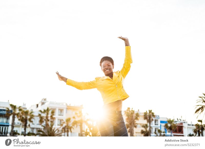 Fröhliche Frau lächelt und springt Glück hüpfen Hände hoch stylisch freudig Afroamerikaner springen Lächeln Jacke hell niedlich attraktiv positiv Gesundheit