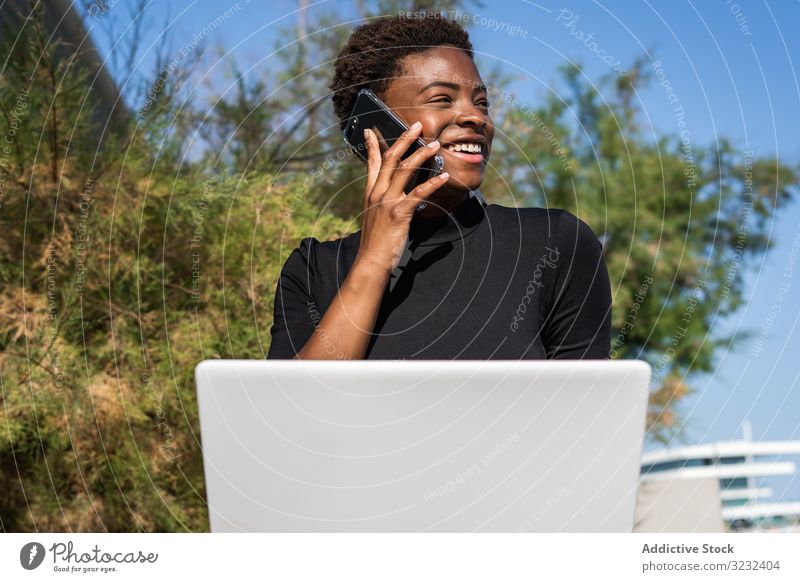 Seriöse Frau surft am Laptop in der Nähe eines modernen Gebäudes sich[Akk] entspannen benutzend Surfen Afroamerikaner elegant Straße Kleid schwarz Straßenbelag