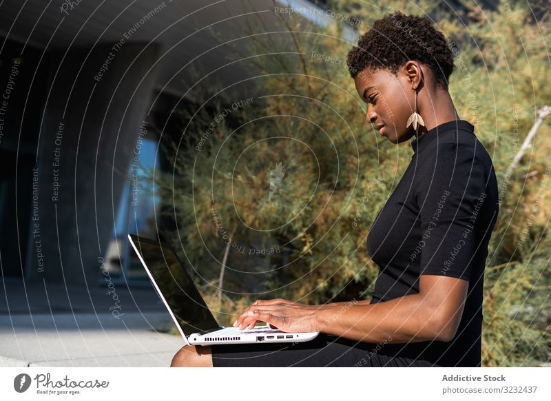 Seriöse Frau surft am Laptop in der Nähe eines modernen Gebäudes sich[Akk] entspannen konzentriert ernst benutzend Surfen Afroamerikaner elegant Straße Kleid