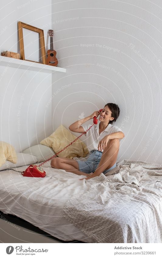 Fröhliche Frau im Telefongespräch Gespräch Lächeln Bett zuhören Dialog Anruf Schlafzimmer Wand sitzen heimwärts Appartement flach retro altehrwürdig Mitteilung