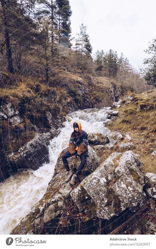 Frau bestaunt schnellen Bergfluss im Wald Berge u. Gebirge Fluss Starrer Blick Strom Sitzen Immergrün Jacke sitzen kalt Herbst Baum reisen Abenteuer Natur