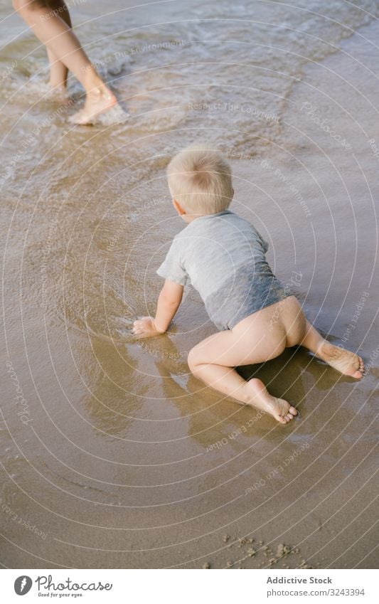 Kleines Kind mit unbekleidetem Hintern krabbelt am nassen Sandstrand Strand Wasser Mutter neugierig tropisch schwimmen auf allen Vieren Kindheit Meer Sohn