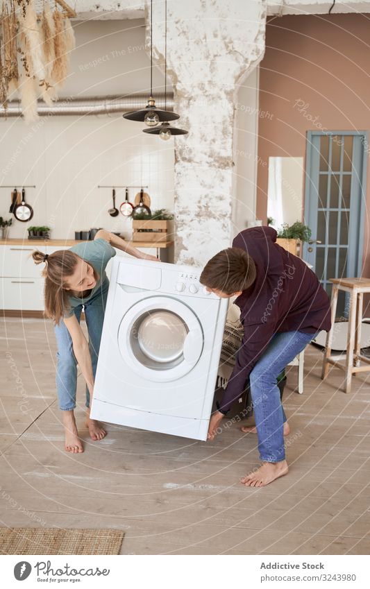 Enthusiastisches Paar trägt Waschmaschine in neue Küche bewegend einrichten Appartement Vorrichtung heimwärts führen heimisch heiter Versand Anwesen