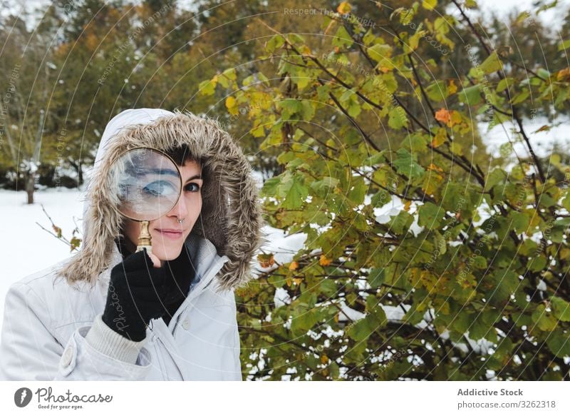 Lächelnde Frau, die im Winter durch eine Lupe schaut interessiert Straße Landschaft Berge u. Gebirge Schnee leer Piercing Inhalt spielerisch weiß neugierig