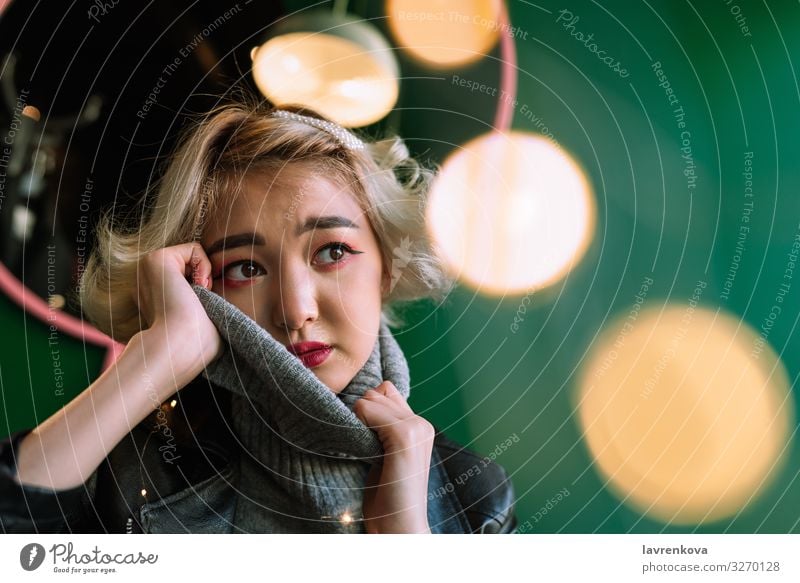 Junge blonde asiatische Frau im gestrickten Rollkragenpullover mit Lichterketten und Bokeh um Weihnachten & Advent Anti-Weihnachten Girlande Asiate attraktiv