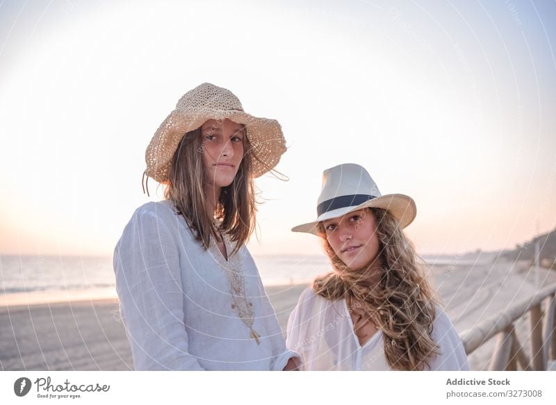 Lächelnde Freundinnen mit Hüten unterhalten sich auf einem Holzzaun am Strand sitzend Frauen MEER reisend Lehnen Seeküste charmant Urlaub Hut lockig Feiertag