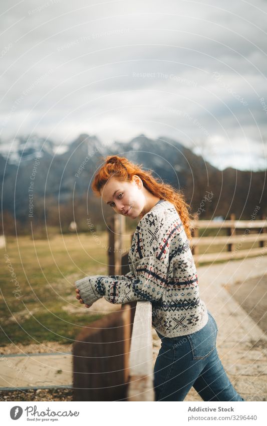 Rothaarige Frau schaut in die Kamera und lehnt an einem Holzzaun entlang einer Straße in den Bergen Lehnen Berge u. Gebirge gemütlich Landschaft warm Pullover