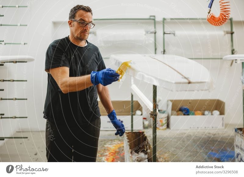 Professioneller Mann malt weißes Surfbrett Kunsthandwerker Kunstgewerbler Holzplatte Herstellung Arbeit Werkstatt Farbe Meister professionell Schutz Handschuhe