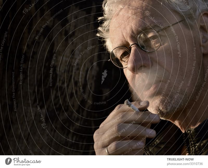 Weißhaariger, rauchender Mann mit Brille und kritischem Blick, brennender Zigarette in der Hand, Ring am kleinen Finger, von der Abendsonne beleuchtet vor dunklem Hintergrund in angeschnittenem Halbprofil untersichtig fotografiert