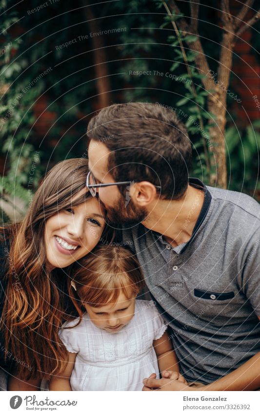 Kleines blondes Mädchen, das zwischen Vater und Mutter sitzt. Vater küsst Mutter auf den Kopf. Die Tochter genießt mit ihren jungen Eltern im Garten. Liebe und Familienkonzept.