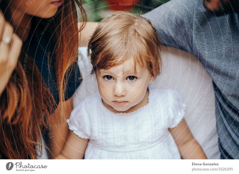 Ernsthaftes kleines blondes Mädchen, das zwischen Mon und Dad sitzt. Gesicht Erholung Kind Baby Kleinkind Eltern Erwachsene Kindheit Kleid sitzen niedlich