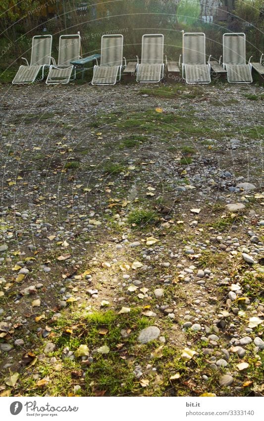 Alte, kaputte, ungepflegte, weisse, leere Liegestühle aus Plastik, stehen nebeneinander in einer Reihe, im Licht auf einem Liegeplatz mit Gras und Steinen und warten auf Urlauber / Besucher.