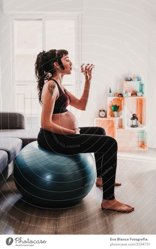 junge schwangere Frau zu Hause, die auf einem Pilates-Ball sitzt und Wasser trinkt. gesunde Lebensweise Yoga heimwärts Sport Gesundheit Lifestyle Jugendliche