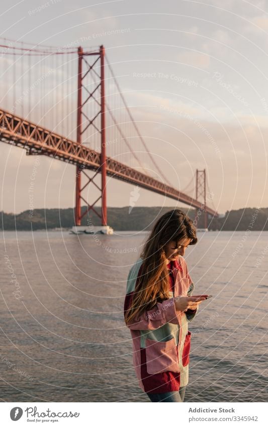 Glückliche Frau surft mit Smartphone gegen Brücke benutzend Handy Nachrichtenübermittlung Freizeit sich[Akk] entspannen Lissabon Portugal Mitteilung Anschluss