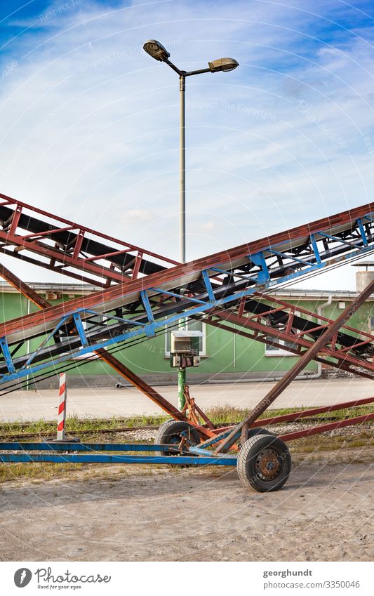 Nebeneinanderstehende Förderbänderwagen, die sich optisch kreuzen. Im Scheitelpunkt ragt eine Straßenlaterne heraus. Auf dem Gelände eines landwirtschaftlichen Betriebs