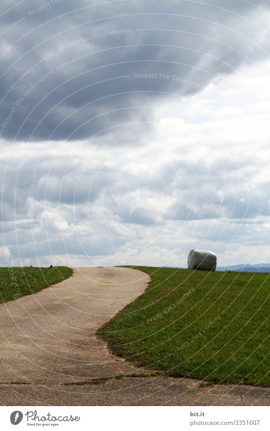 Eingepackter Heuballen liegt nach der Ernte vom Bauer, in der Landwirtschaft, auf der Wiese vom Feld; Acker in einer Landschaft mit Wolken am Himmel und einem Feldweg; Straße mit Kurve.