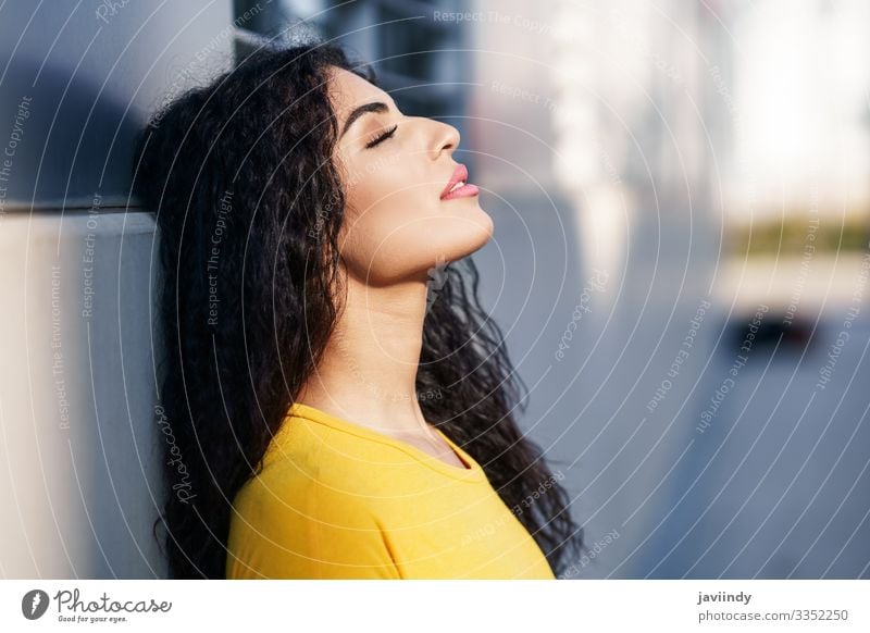 Junge arabische Frau mit geschlossenen Augen und mit dem Kopf an die Wand gelehnt auf der Straße. Lifestyle Stil schön Haare & Frisuren Schminke Sonne Mensch