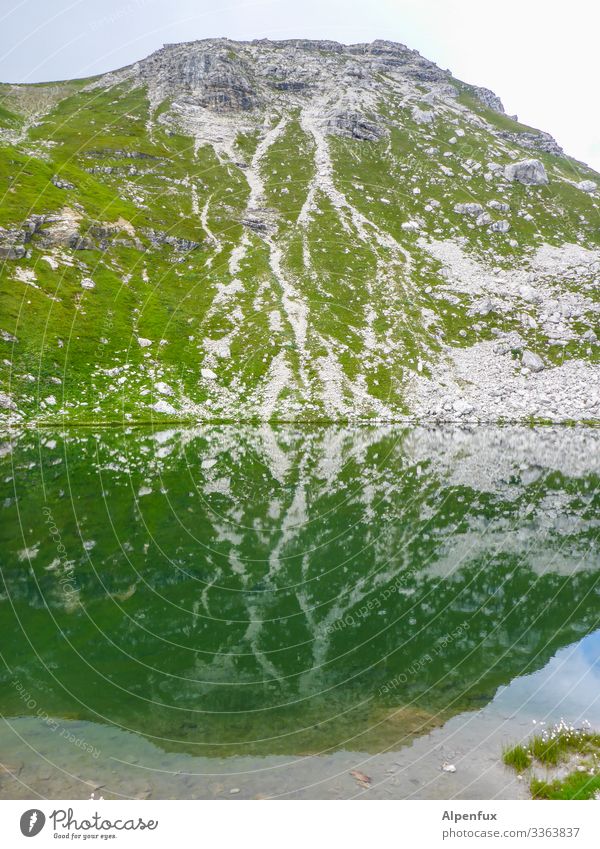 Ein mit Gras bewachsener und mit mäandernden hellgrauen Schotterrinnen nicht sehr hoher unscheinbarer menschenleerer allgäuer Hügel der aussieht wie ein Vulkan spiegelt sich ein paar Farbnuancen dunkler und unscharf in einem kristallklaren Bergsee wieder