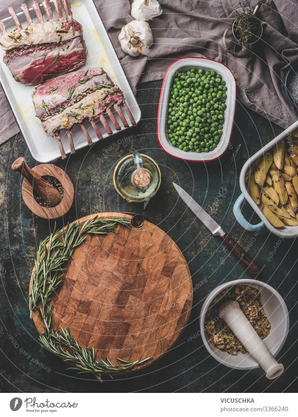 Hintergrund zum Thema Fleischwaren. Kochzubereitung mit rohem Lammfleisch, grünen Erbsen, Backkartoffeln und Rosmarinbrotkruste auf rustikalem Tisch mit leerem Schneidebrett. Ansicht von oben. Lamm-Karren