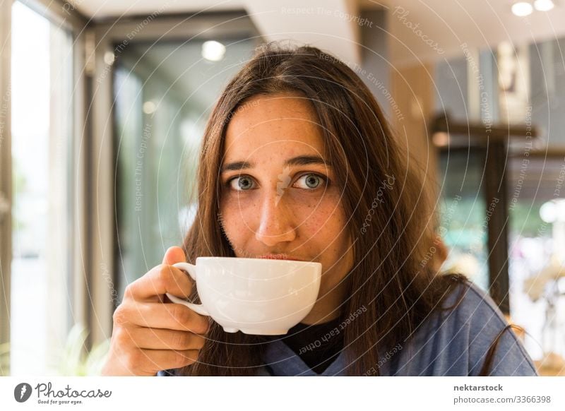 Junge Frau mit Kaffeetasse auf den Lippen schaut in die Kamera Porträt gebräunte Haut hell gefärbte Augen Tasse Becher trinken Getränk Porzellan