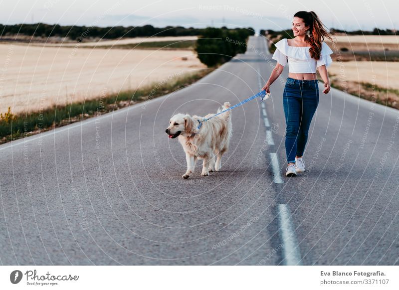 eine junge Frau, die bei Sonnenuntergang mit ihrem Golden Retriever-Hund an der Straße spazieren geht. Haustiere im Freien urban Landschaft Feld laufen Glück