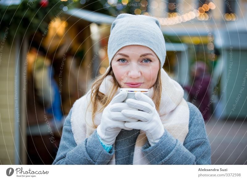 Junge Frau auf dem Weihnachtsmarkt trinkt eine Tasse heiße Schokolade mit Marshmallow und trägt eine warme Strickmütze und einen Schal. Beleuchtete und dekorierte Kioske und Geschäfte im Hintergrund. Helsinki, Finnland