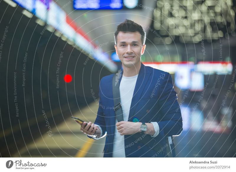 Junger, stilvoller, gut aussehender Mann im Anzug mit Koffer steht auf der Metrostation, hält ein Smartphone in der Hand, scrollt und simst, lächelt und lacht.