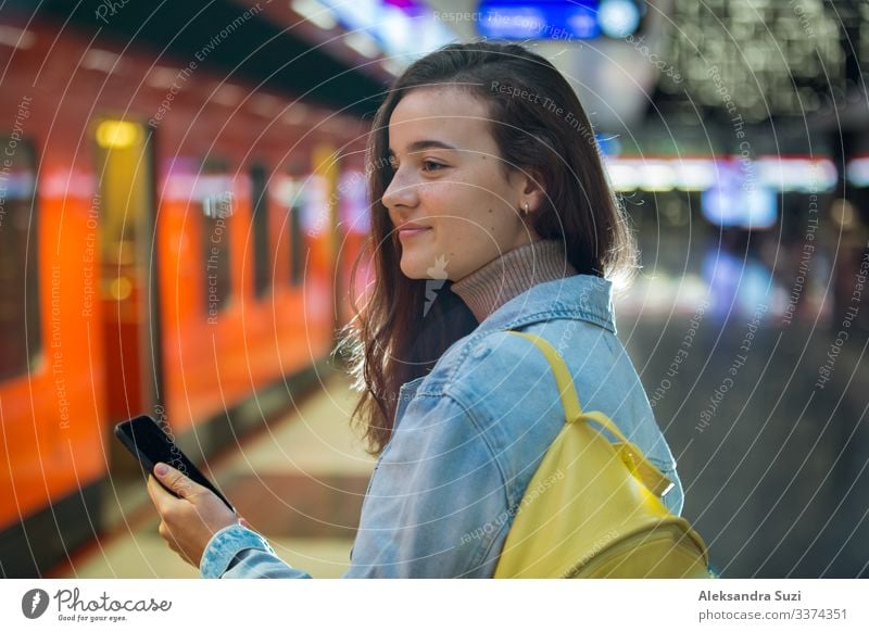 Teenager-Mädchen in Jeans mit Rucksack steht in der U-Bahn-Station, hält Smartphone in der Hand, scrollt und schreibt SMS, lächelt und lacht. Futuristisch helle U-Bahn-Station. Finnland, Espoo