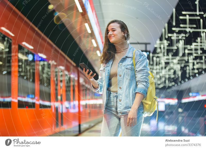 Teenager-Mädchen in Jeans mit Rucksack steht in der U-Bahn-Station, hält Smartphone in der Hand, scrollt und schreibt SMS, lächelt und lacht. Futuristisch helle U-Bahn-Station. Finnland, Espoo