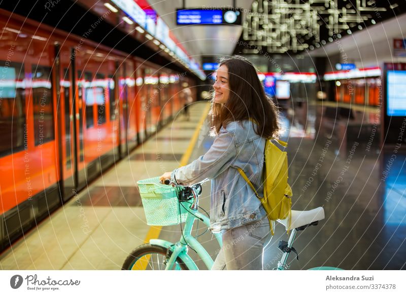 Teenager mit Rucksack und Fahrrad auf der U-Bahn-Station stehend, Smartphone in der Hand haltend, blätternd und simsend, lächelnd und lachend. Futuristisch helle U-Bahn-Station. Finnland, Espoo