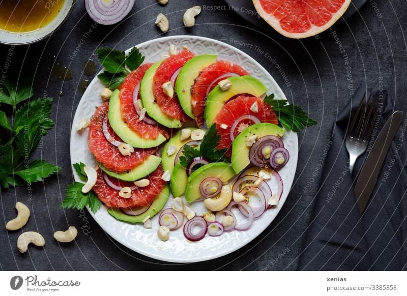 Salat mit Avocado, Grapefruit, Zwiebeln, Cashewnüsse und Petersilie auf weißem Teller angerichtet mit Messer und Gabel in schwarzer Serviette auf dunkler Tischplatte