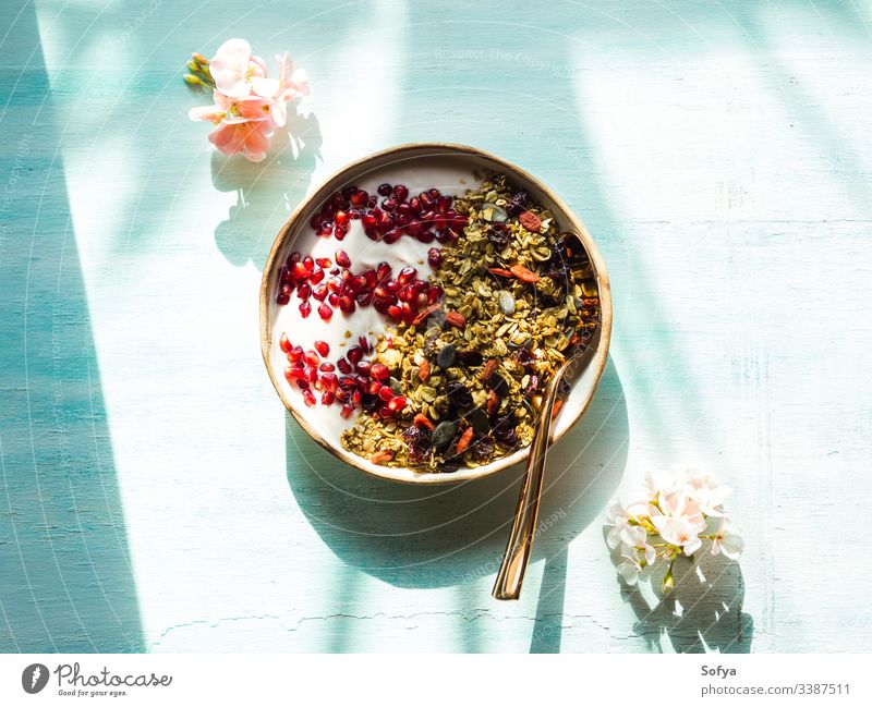 Hausgemachte Müslimaschüssel mit Granatapfelkernen, Matcha-Müsli mit Hafer und Trockenfrüchten, Nüsse auf türkisem Hintergrund mit rosa Blüten. Frühstück lecker