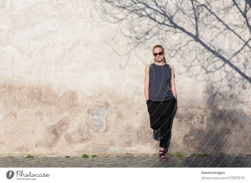 Grafisches und texturiertes künstlerisches Bild einer modernen, modischen Frau mit Sonnenbrille, die sich an eine alte, texturierte Retro-Wand lehnt, wobei der Schatten eines Baumes auf die Wand fällt.