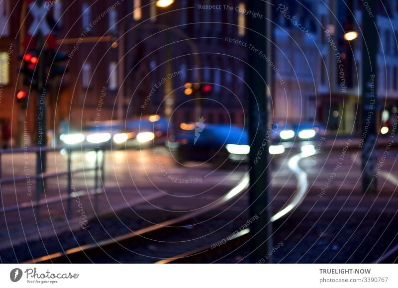 Straßenkreuzung am Abend mit Ampeln, Autos, die mit Licht fahren und elegant gebogene Tram-Schienen, die das Scheinwerferlicht reflektieren. Im Hintergrund ein großes Eckhaus aus dunkelroten Backsteinen.