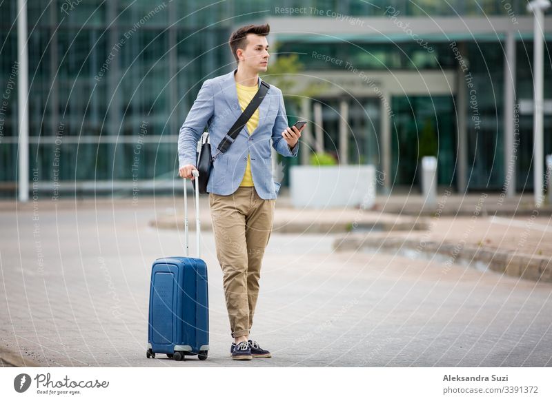 Stylischer Mann steht am Flughafen mit Smartphone und Koffer, blättert, textet, benutzt mobile Apps. Geschäftsreisende. Anwendung Ankunft Tasche Business